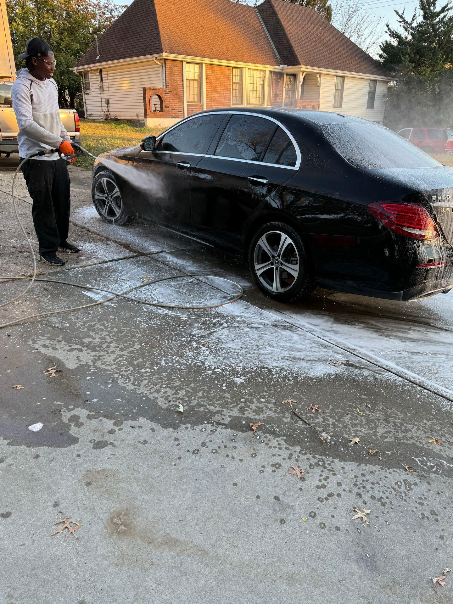 Person washing a black car with a high-pressure water hose on a driveway near a house.