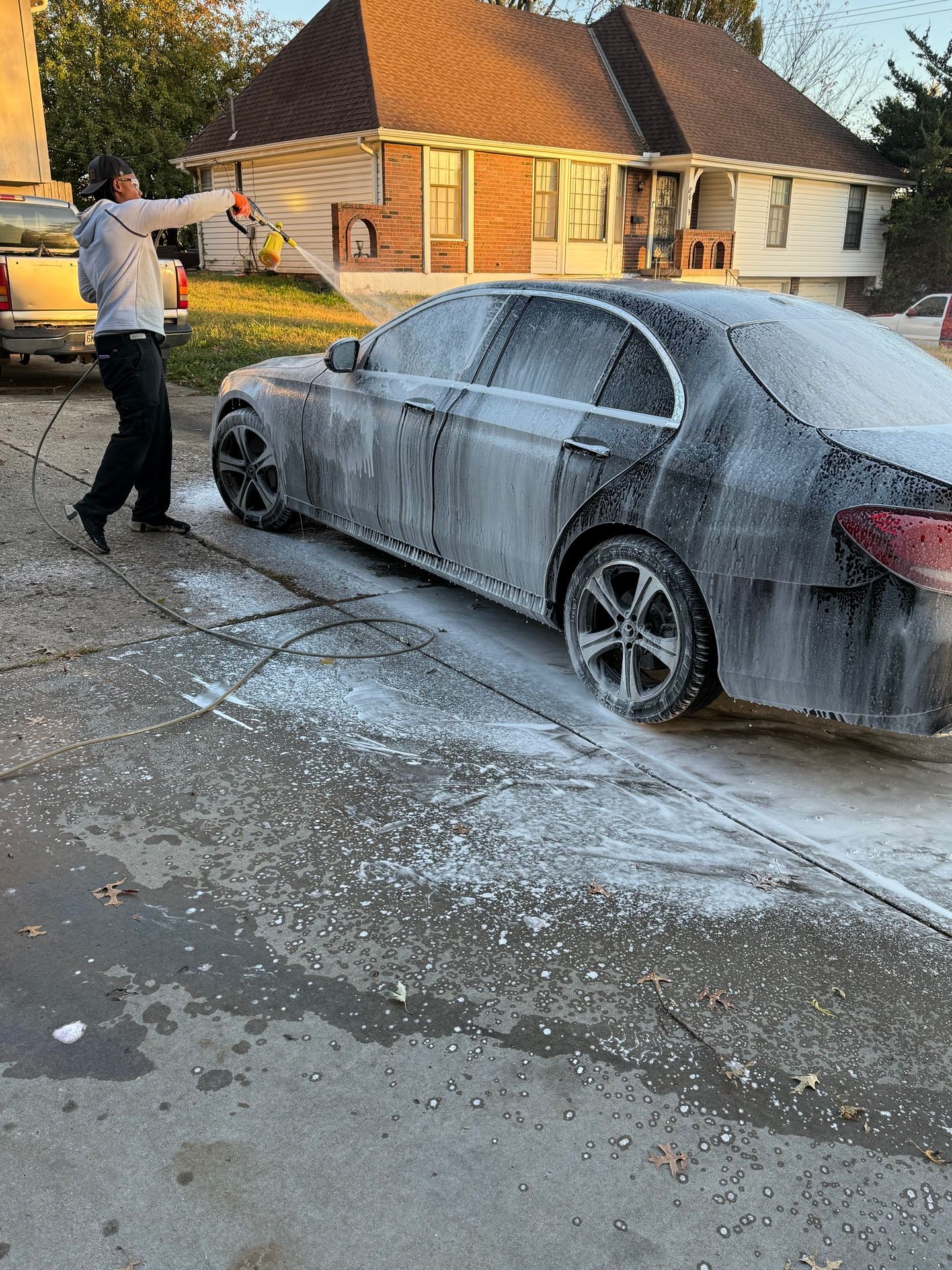 Person washing a car covered in soap foam near a residential house on a sunny day.