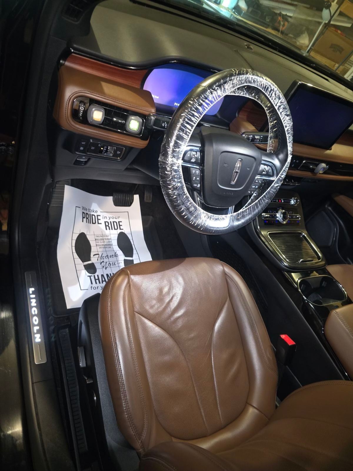 Interior of a luxury car with brown leather seats and plastic-wrapped steering wheel.